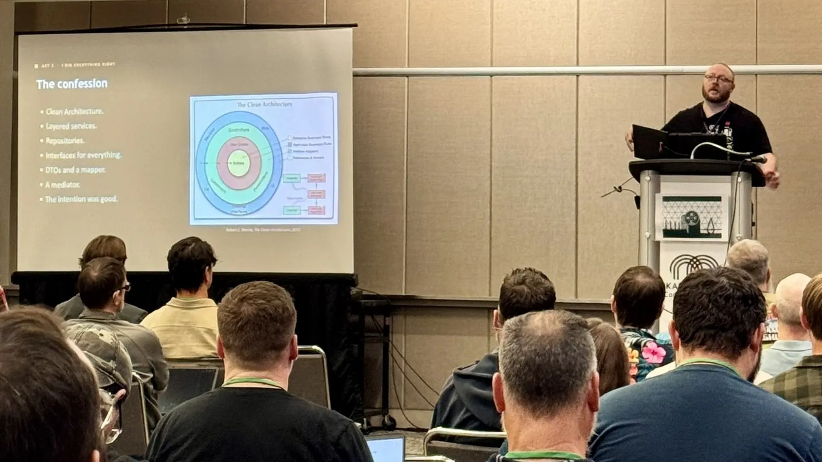 Erik Shafer speaking from the podium at KCDC 2026, beside a slide titled "The confession" that shows the Clean Architecture diagram, with a full room in the foreground.